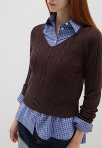 Maglione a coste marrone con un motivo a trecce su una camicia a scacchi blu con bottoni. Il maglione presenta un colletto a V e maniche lunghe.