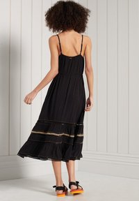 Svart maxiklänning med tunna axelband, rynkat liv, lager på nederdelen och detaljer i guldbroderi. Bärs med svarta sandaler med orange sulor.