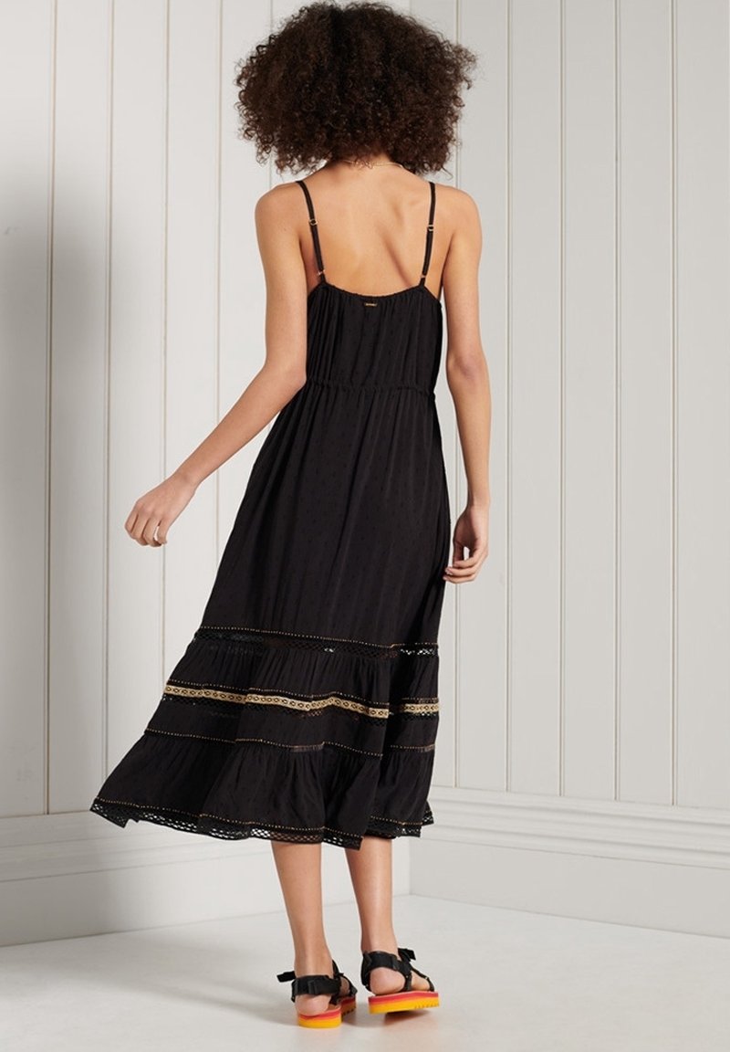 Svart maxiklänning med tunna axelband, rynkat liv, lager på nederdelen och detaljer i guldbroderi. Bärs med svarta sandaler med orange sulor.