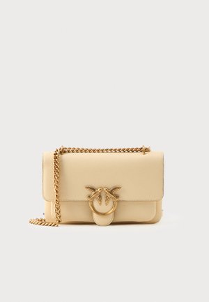 Krémová kožená crossbody taška se zlatým řetízkem a dekorativním zlatým zapínáním ve tvaru prstenu, na kterém jsou dvě ptáky čelící si navzájem.