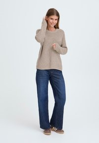 Beige gestrickter Pullover mit weitem Ausschnitt und langen Ärmeln, kombiniert mit dunkelblauen weiten Jeans und braunen Schuhen mit weißen Streifen.