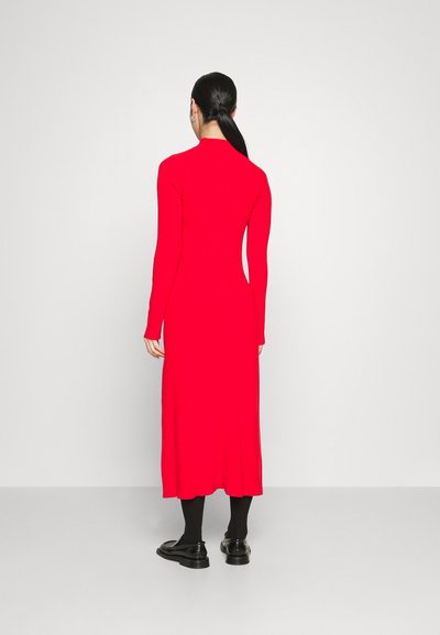 Robe rouge à manches longues avec une silhouette ajustée, ourlet droit et texture en tricot, associée à des collants et des chaussures noires.