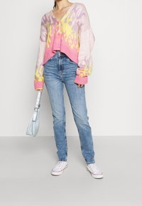 Rosa och gula flammönstrade kortärmad cardigan med V-ringning, knäppning och ribbade ärmslut, kombinerad med ljusblå jeans och vita sneakers.