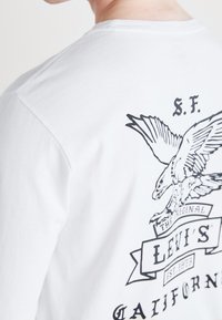 Weißes Langarm-T-Shirt mit einem schwarzen Grafikdruck eines Adlers und Text auf dem Rücken, mit rundem Halsausschnitt und weichem Baumwollmaterial.