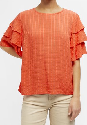 Nainen, joka pukeutunut korallinoranssiin ruudulliseen puseroon, jossa on kerrokselliset röyhelöhihat, yhdistettynä beigeruskeisiin housuihin.