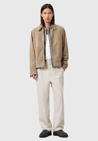 Tan suede jacket with a pointed collar, layered over a checkered shirt, paired with beige tailored trousers and dark shoes.