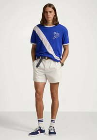 Young man wearing blue Polo shirt with white diagonal stripe, white shorts, striped belt, white socks, and blue sneakers standing against plain background.
