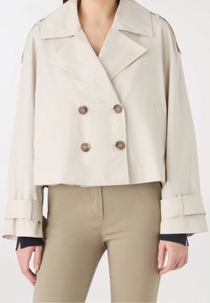Donna che indossa una giacca corta doppiopetto beige con ampi revers e polsini con cintura sopra pantaloni beige.