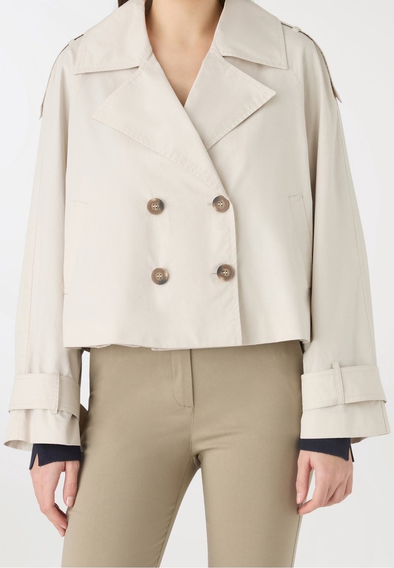 Femme portant une veste courte beige à double boutonnage avec de larges revers et des manches ceinturées sur un pantalon beige.
