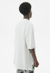 Přehozené bílé tričko s krátkým rukávem a kulatým výstřihem, měkkou texturou a přírodními detaily na lemu. Kombinováno se vzorovanými černými šortkami.