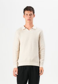 Beige strikkegenser med teksturfinish, V-hals og kontrastkrage. Skulderpassede ermer og ribbestrikket kant. Båret over mørke bukser.