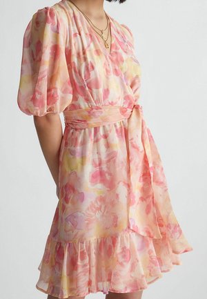 Kobieta w pastelowej, kwiecistej sukience kopertowej z bufiastymi rękawami, przewiązanym paskiem w talii oraz warstwowym falbanowym dołem, ozdobiona warstwowymi złotymi naszyjnikami.