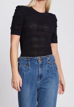 Femme portant un haut en maille noir à manches courtes avec des motifs texturés et un jean bleu taille haute avec des détails de boutons à la taille.