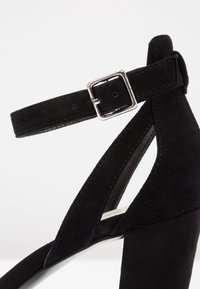 Chaussure à talon haut en daim noir avec bride cheville et boucle argentée, avec des côtés découpés et un talon bloc robuste.