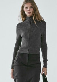 Grauer gerippter Strickpullover mit hohem Kragen und Frontreißverschluss, mit langen Ärmeln und figurbetontem Schnitt, kombiniert mit einem dunklen Rock.