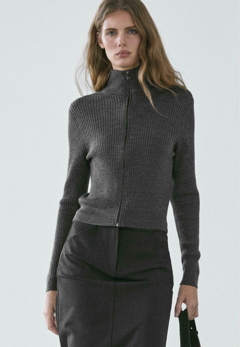 Grauer gerippter Strickpullover mit hohem Kragen und Frontreißverschluss, mit langen Ärmeln und figurbetontem Schnitt, kombiniert mit einem dunklen Rock.