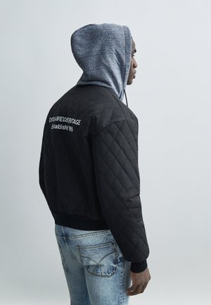 Homme portant un sweat à capuche gris sous une veste matelassée noire avec un texte blanc au dos, associé à un jean bleu clair délavé, faisant face à droite sur un fond uni.