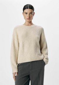 Pull en laine beige clair avec encolure ronde, poignets et ourlet côtelés, texture douce et coupe décontractée, associé à un pantalon gris.
