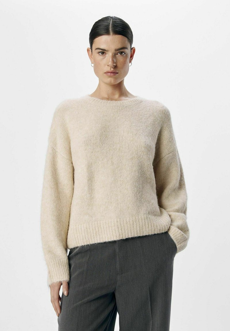 Suéter de lana color beige claro con un cuello redondo, puños y dobladillo acanalados, textura suave y corte relajado, combinado con pantalones grises.