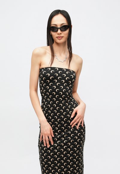 Czarna sukienka bez ramiączek z kremowymi wzorami w kształcie półksiężyców, wykonana z gładkiego materiału. Dopełniona srebrnym naszyjnikiem i czarnymi okularami przeciwsłonecznymi.