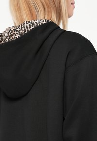 Svart hettegenser med leopardmønstret fôr på hetta. Myk tekstur med løs passform og synlige sømdetaljer på skulderen.