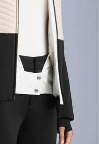 Kadar izbliza osobe koja nosi crno-bijelu jaknu sa zipom i dugmetima na drukere, preko crnih hlača i bijele majice.