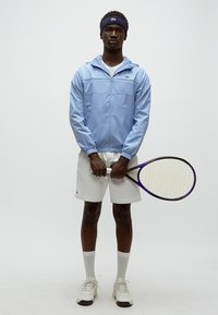 Casaco azul claro com fecho de correr e punhos elásticos; calções brancos; a segurar uma raquete de ténis roxa e preta; meias e sapatos brancos.