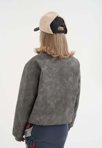 Giacca in suede grigio con design ricamato sul retro, indossata con un cappello beige e scuro, caratterizzato da lunghi capelli biondi e una gonna a fantasia.