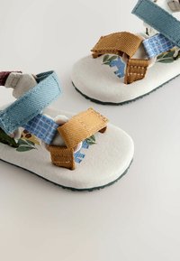Sandales en tissu pour enfants avec brides réglables bleues et beige, et motif floral et dinosaure imprimé sur la semelle.