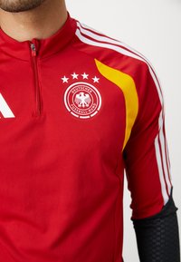 Röd sporttröja med hög krage, som har vita ränder på ärmarna, en gul accent och en rund logotyp med en dubbelhövdad örn och stjärnor.