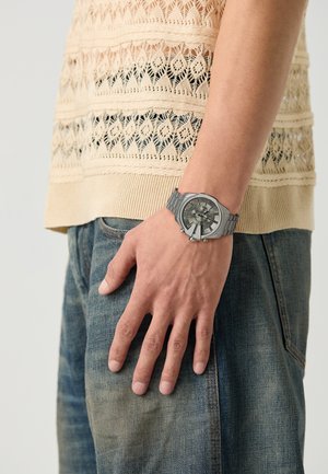 Montre bracelet en argent avec un cadran rond, un bracelet métallique et des cadrans détaillés. Portée sur une main reposant sur un jean en denim, contre un haut en beige texturé.