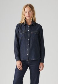 Camisa de ganga escura com frente abotoada, dois bolsos no peito e botões brancos. O tecido tem uma textura suave com costura em contraste.