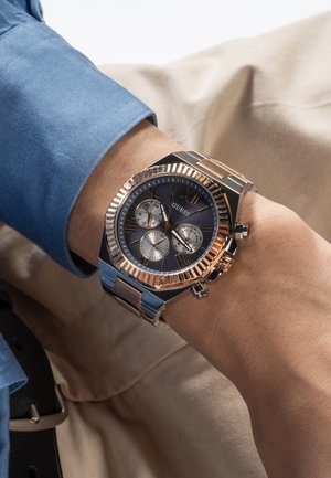 Ročna ura z črno-zlatim kovinskim pasom na levi zapestju, ki prikazuje čas, datum in dan, nošena z modro srajco in bež hlačami.