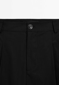 Pantalon noir avec une fermeture à bouton à la taille, présentant des plis et des coutures nettes. Le tissu semble lisse avec une finition mate.