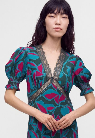 Femme aux longs cheveux foncés portant une robe fleurie turquoise et magenta avec des manches bouffantes et une bordure en dentelle noire, debout devant un fond uni.
