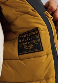 Interior de chaqueta aislante amarilla y negra con textura acolchada, que presenta una etiqueta con "HAWKDOWN" y detalles de marca, además de un zipper amarillo.