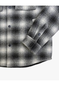 Camicia a quadri nei colori nero, grigio e bianco. Realizzata in tessuto morbido con un motivo houndstooth. Presenta bottoni neri e un orlo arrotondato.