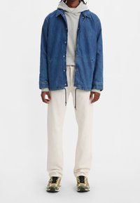 Chaqueta de mezclilla azul superpuesta a una sudadera gris, combinada con pantalones color crema y zapatillas multicolores con cordones y detalles negros.