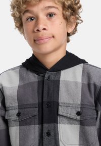 Černá a šedá kostkovaná flanelová košile s černou kapucí, se dvěma náprsními kapsami a černými knoflíky, měkká textura a ležérní design.