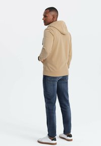Sudadera beige con cordón, mangas largas y bolsillo tipo canguro, combinada con vaqueros azul oscuro y zapatillas blancas con detalles negros.