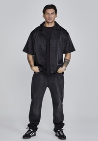 Camisa negra de manga corta con un diseño texturizado, combinada con una camiseta negra, jeans de mezclilla oscuros y zapatillas negras con rayas blancas.