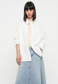 Hvit knapp-up skjorte, lett stoff, oversized passform, lange ermer, enkelt brystlomme. Kombinert med en lys blå denimskjørt.