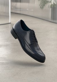 Black polished leather dress shoe with lace-up design floating above light gray concrete floor in modern indoor setting.