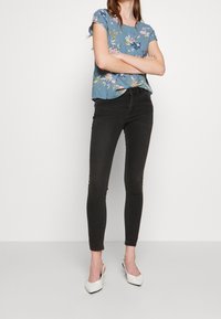 Top de estampado floral en azul con mangas cortas, combinado con jeans skinny negros y tacones blancos de punta. Tejido texturizado con un corte holgado.