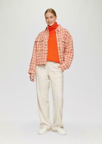 Rot-weiße Hahnentrittmuster-Jacke, orangefarbener Rollkragenpullover, cremefarbene weit geschnittene Hose und weiße Sneaker. Die Jacke hat zwei vordere Taschen.