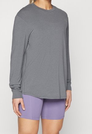 Personne portant une chemise grise ample à manches longues et des shorts de cyclisme lavande ajustés, les mains détendues le long du corps.