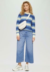Gestrickter Pullover mit blauen und weißen Streifen, weit geschnittene hellblaue Jeans und eine cremefarbene Umhängetasche. Die Schuhe sind graue Sneakers.