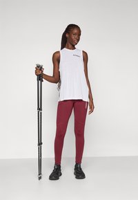 Top bianco senza maniche con logo nero, leggings bordeaux con dettagli, e bastoni da escursionismo neri. Texture liscia, vestibilità atletica e design funzionale.