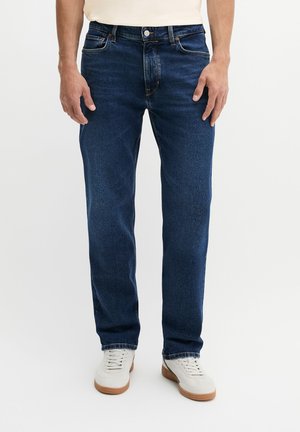 Mann trägt dunkelblaue gerade geschnittene Jeans und weiße Sneakers und steht vor einem schlichten hellen Hintergrund.