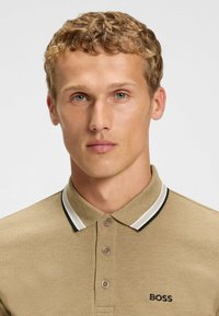Khakifarbenes Poloshirt aus strukturiertem Material mit einem Zwei-Knopf-Feplaintext, kontrastierenden weißen und schwarzen Kragestreifen sowie einem gestickten "BOSS"-Logo.